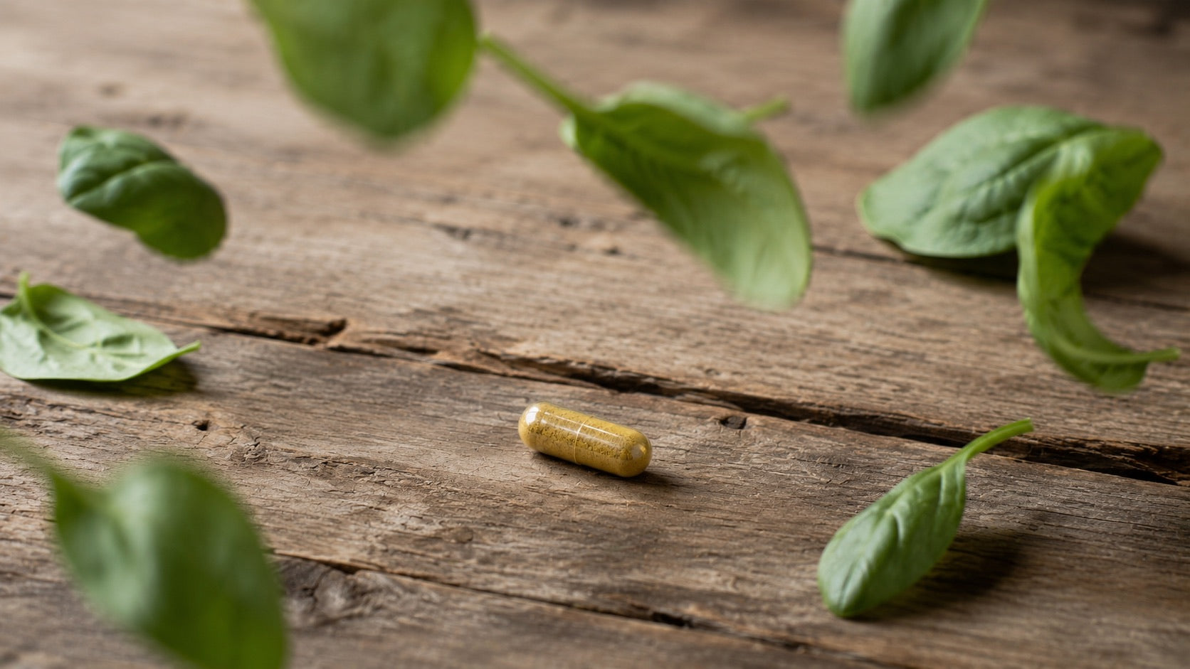 Transparente Multivitamin-Kapsel mit senffarbenem Pulver auf Altholz, während Spinatblätter darüber herabfallen.