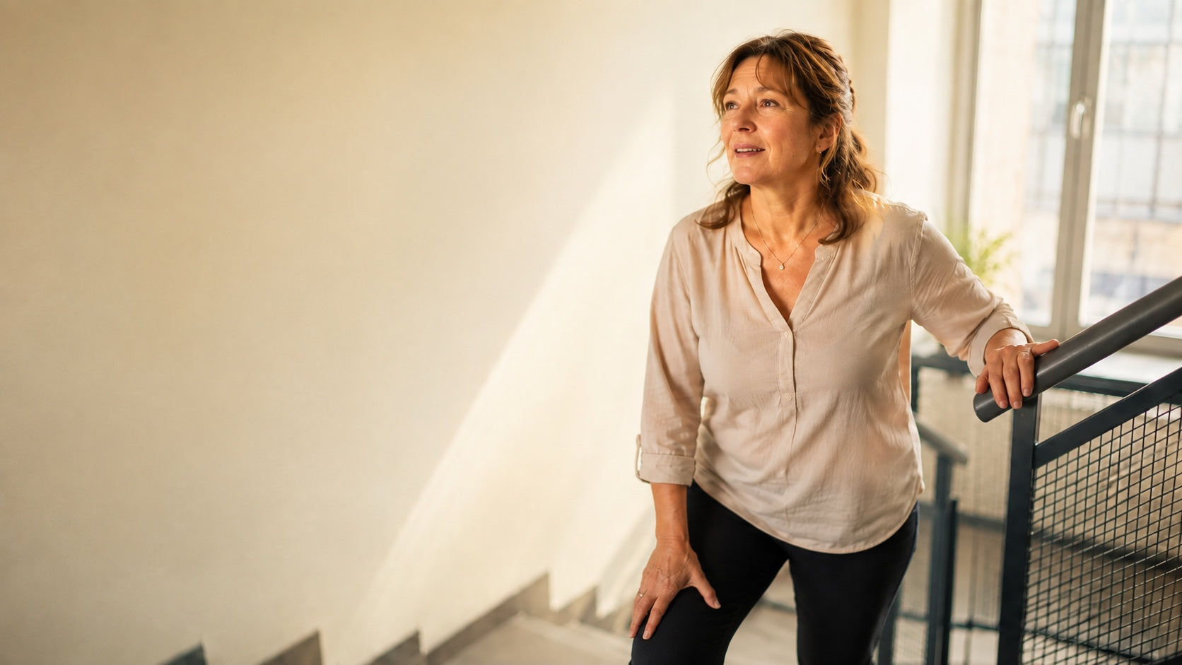 Birgit, 54, steht in einem hellen Treppenhaus und hält kurz inne, nachdem ihr das Treppensteigen zunehmend Kraft kostet.