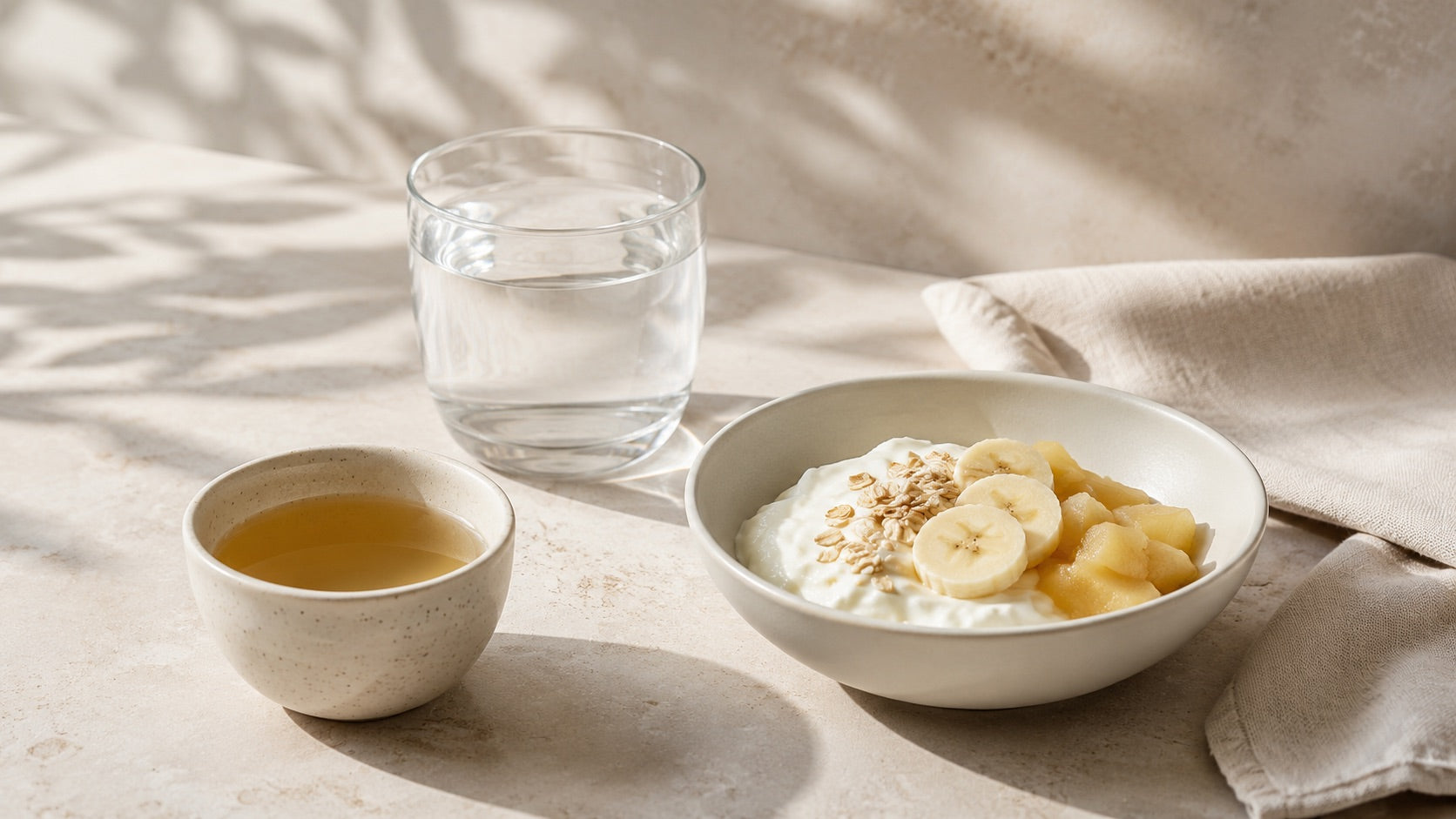 Leicht verdauliche Mahlzeit mit Joghurt, Banane, Apfel und Haferflocken neben Wasser und Tee auf einem hellen Tisch.