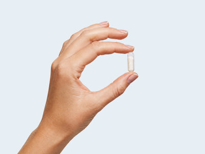 Weiße CALM Magnesium-Kapsel von Fifty Five zwischen Daumen und Zeigefinger vor hellem Hintergrund