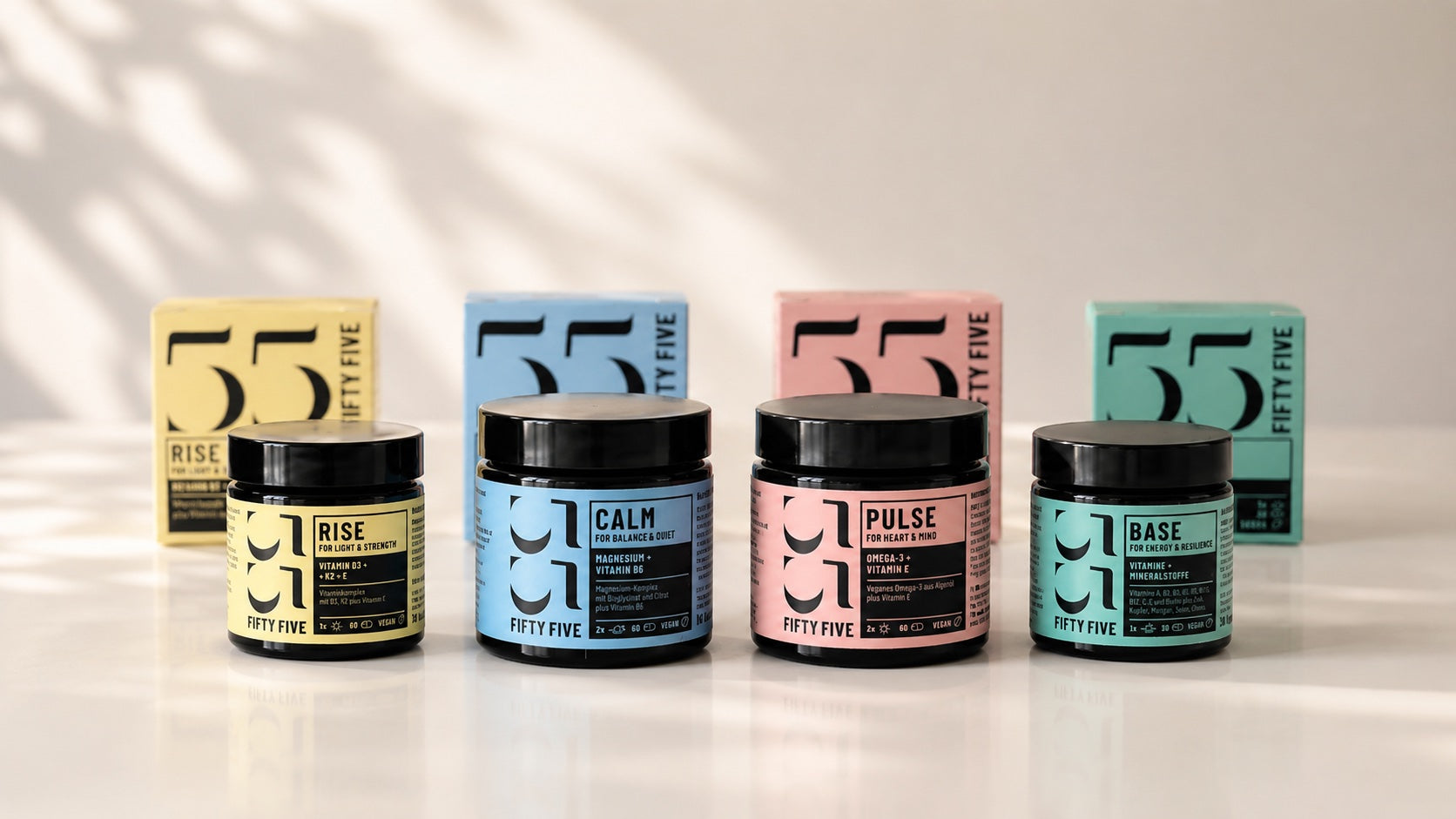 Vier Fifty Five Daily Essentials Produkte mit passenden Boxen im Hintergrund auf heller Oberfläche: RISE, CALM, PULSE und BASE in natürlichem Studiolicht.