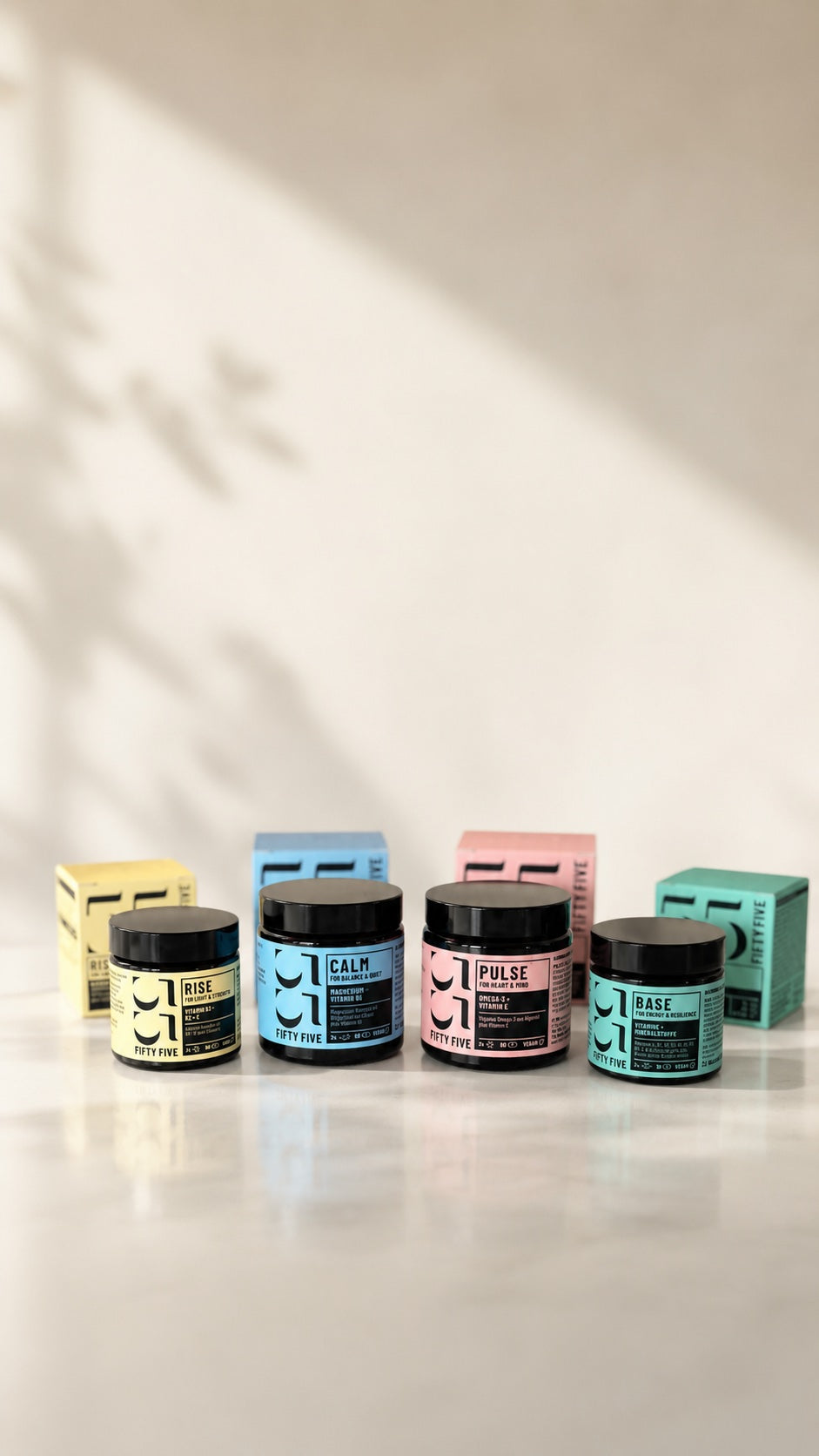 Vier Fifty Five Daily Essentials Produkte mit passenden Boxen im Hintergrund auf heller Oberfläche: RISE, CALM, PULSE und BASE in natürlichem Studiolicht.