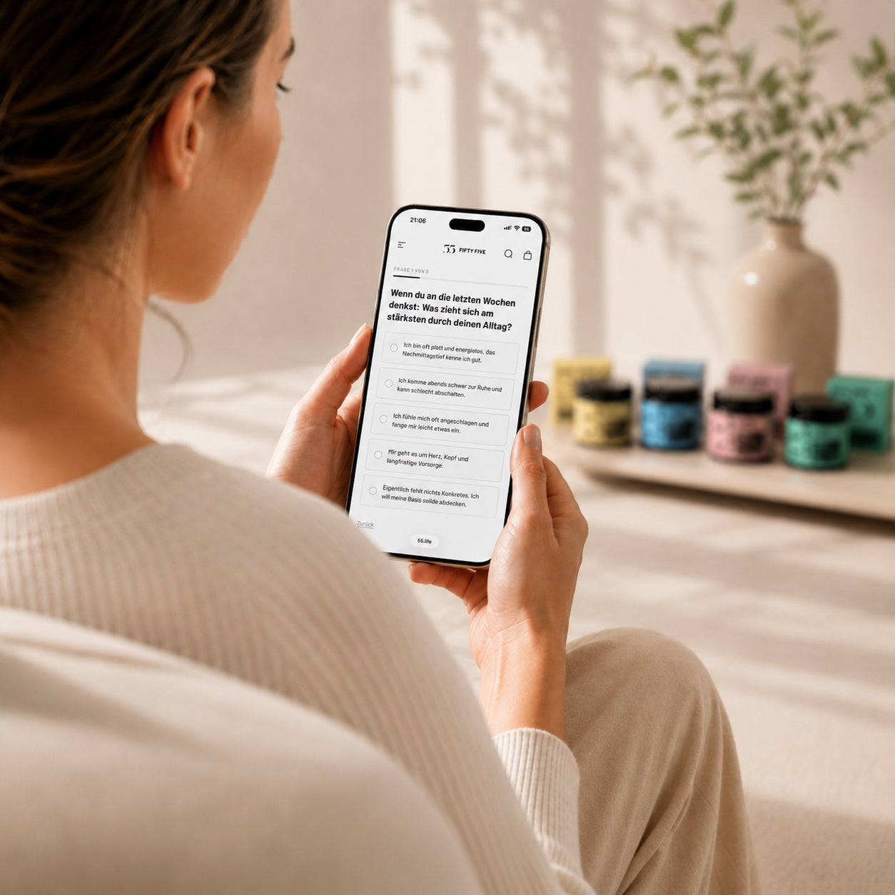 Nahes Lifestyle-Motiv einer Frau mit iPhone beim Fifty Five Bedarfs-Check, mit weich unscharfen Supplement-Produkten im Hintergrund.