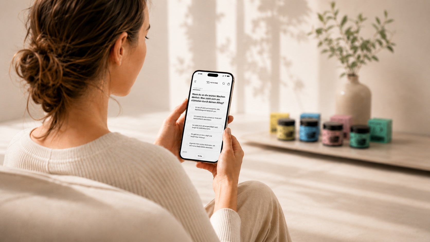Frau hält ein iPhone mit dem Fifty Five Bedarfs-Check in den Händen, während Fifty Five Produkte im Hintergrund leicht unscharf auf einer hellen Oberfläche stehen.